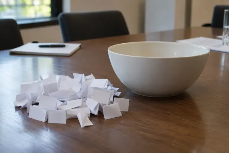 Identical folded paper slips beside a plain bowl on a meeting table.