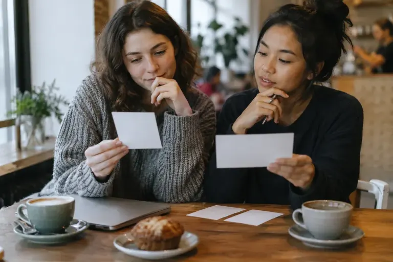 Two friends discussing a choice using blank decision cards at a cafe table, thoughtful ordinary scene.